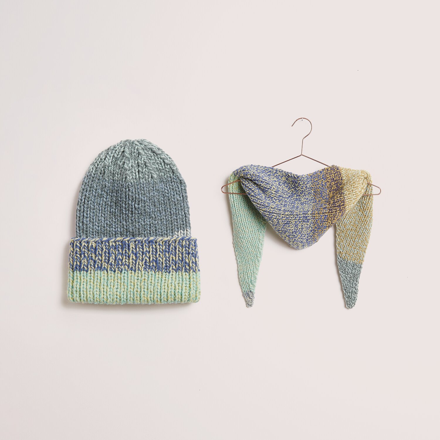 Die Neue Masche No. 14 Winter von idee. Creativmarkt Strickset Mütze + Dreieckstuch Modell 22 aus DNM 14 in sanften Pastelltönen, präsentiert auf einem Kleiderbügel. Ideal für den Winter.