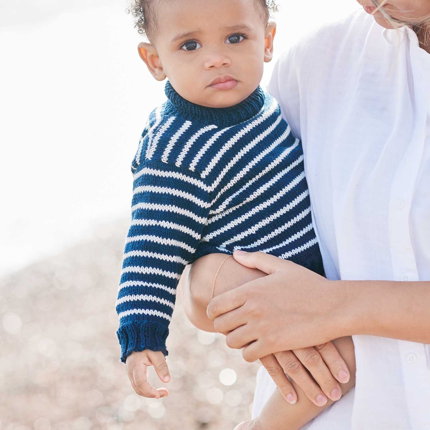 Strickanleitung Pullover aus Baby Organic Cotton: Ein Baby trägt einen blau-weiß gestreiften Pullover am Strand, gehalten von einer Person in weißem Hemd.