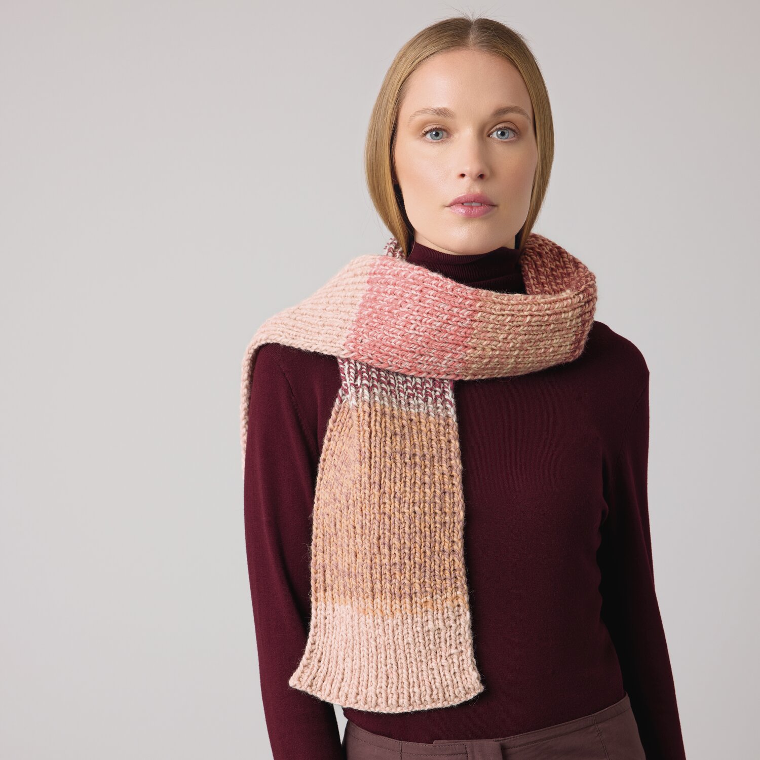Strickset Schal Modell 04 aus Die Neue Masche No. 14 von idee. Creativmarkt Strickset Schal Modell 04 aus DNM 14: Frau trägt gestrickten Schal in Rosa- und Beigetönen über einem weinroten Pullover.