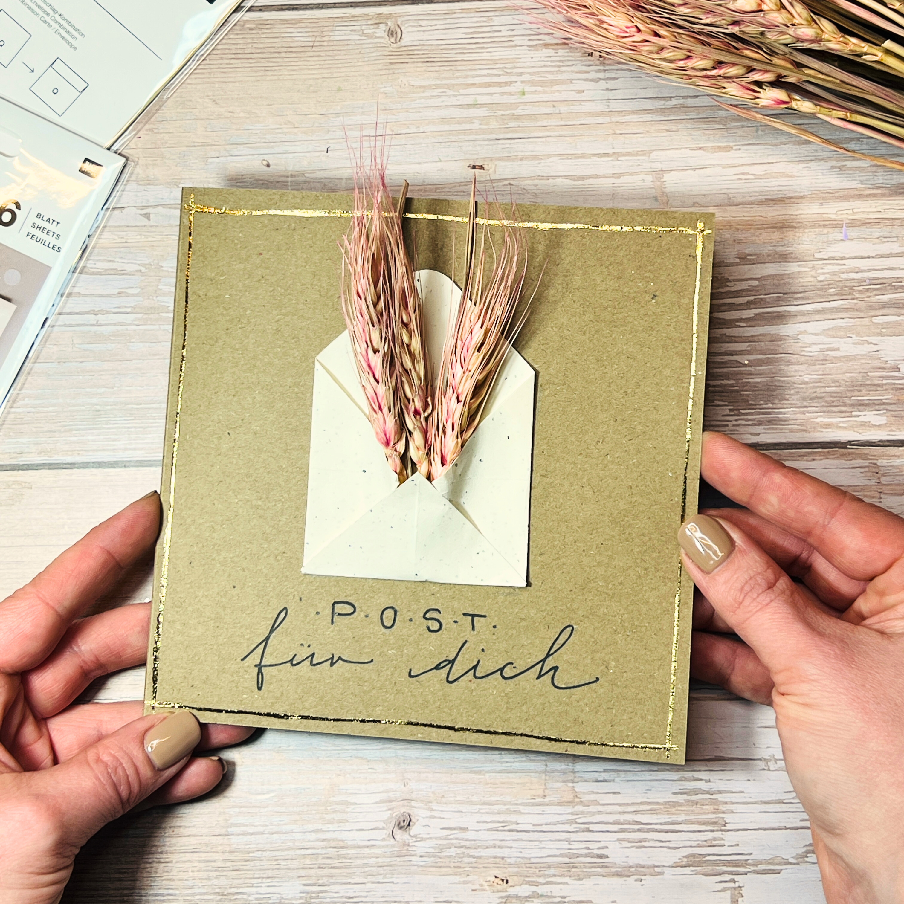Eine quadratische Karte, mit weißem Mini-Umschlag und Trockenblumen, sowie einem Schriftzug, wird von Händen gehalten.