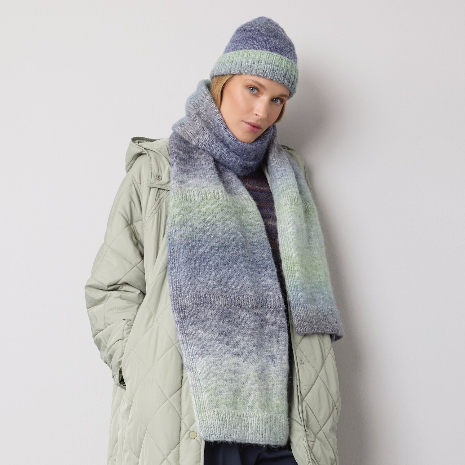 Strickset Mütze Modell 20 aus Die Neue Masche No. 14 von idee. Creativmarkt Strickset Mütze Modell 20 aus DNM 14: Frau trägt gestrickte Mütze und Schal in Blau- und Grüntönen, kombiniert mit beigem Mantel.