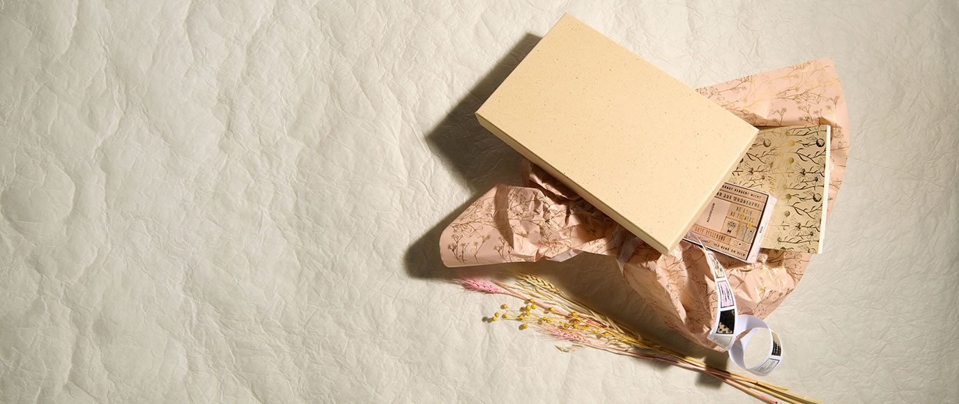 Geöffnete Geschenkbox mit dekorativem Seidenpapier, Papierprodukten und Trockenblumen auf hellem Hintergrund.