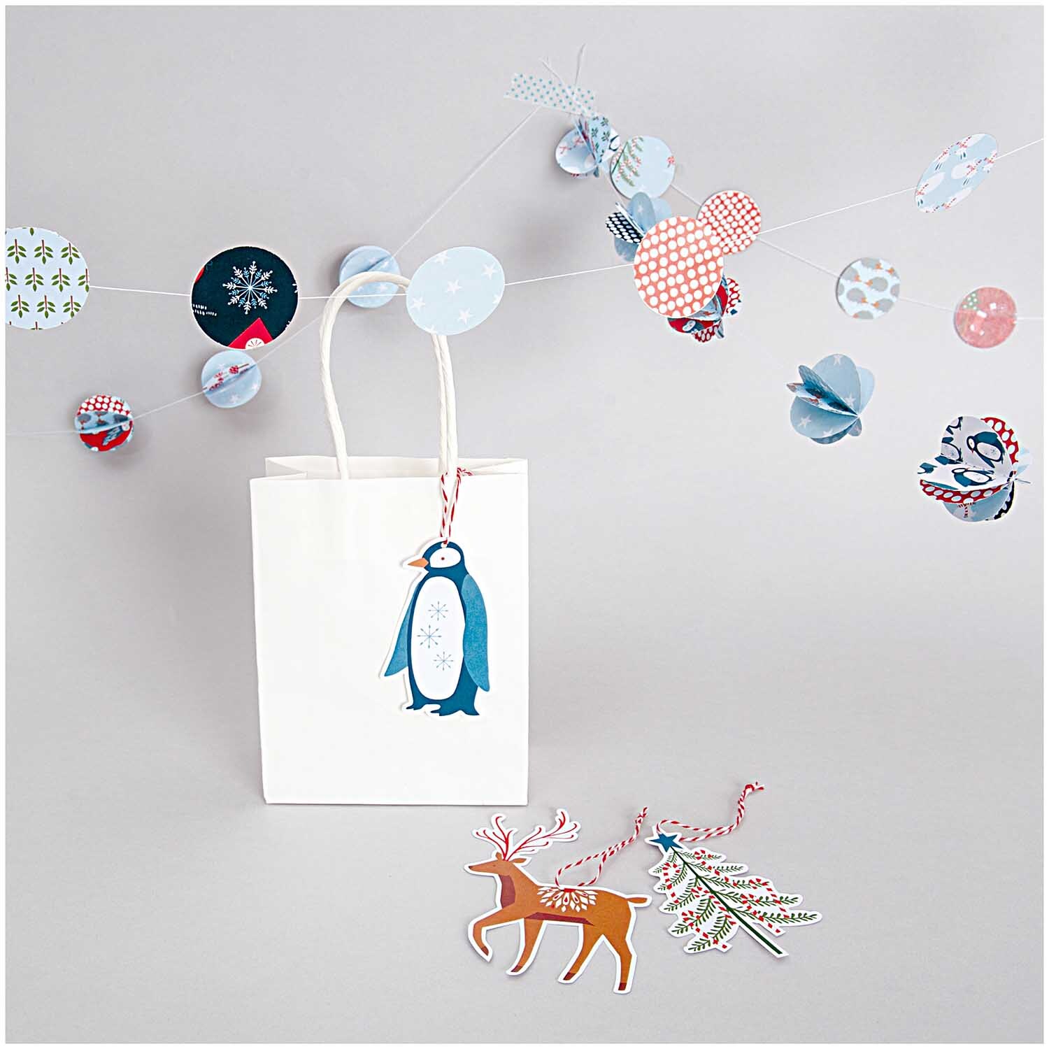 Papiertüte weiß von Rico Design mit Pinguinmotiv, umgeben von bunten Papierdekorationen.